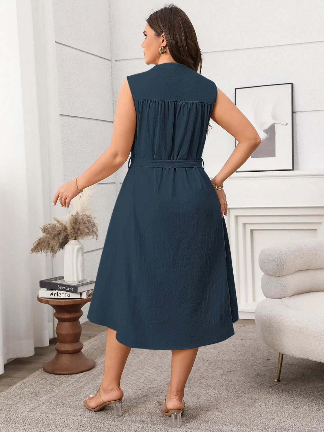 Woman wearing sleeveless tie waist plus size dress in navy blue standing in modern living room
